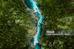 Drone high-angle photo of the turquoise-colored mountain river flowing in the pine woodland with a view of the mountain peaks in the background in Innlandet County, Norway